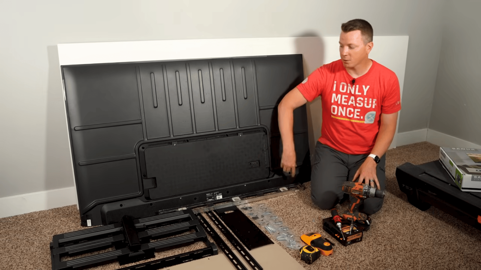 Person preparing a TV for wall mounting with a bracket, drill, screws, and tools on the floor.