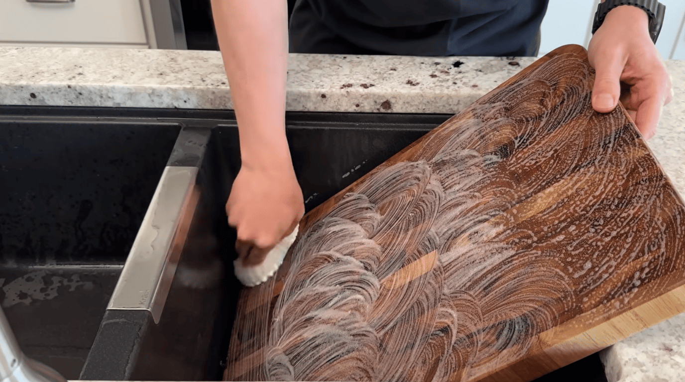 person washing a wooden cutting board with soap and sponge near a kitchen sink for proper cleaning