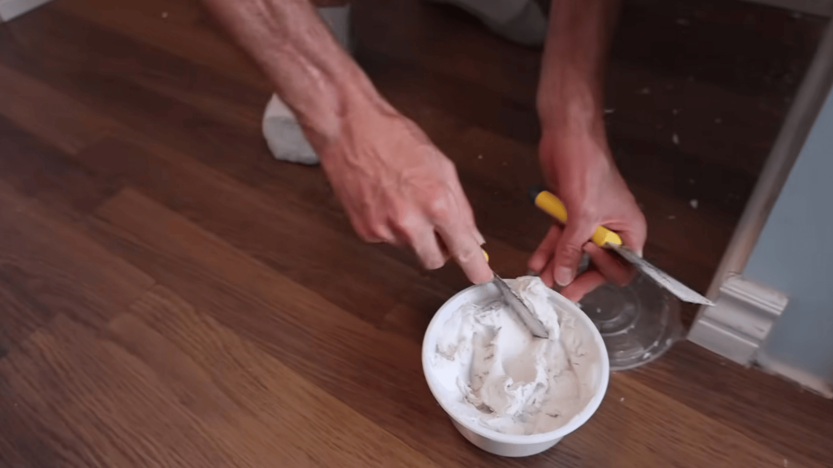 Hands mixing drywall mud in a bowl with a putty knife for wall repair work.