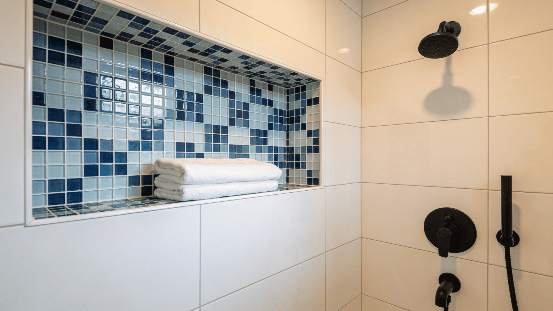 Shower niche with blue mosaic tile and folded towels, beside a matte black rain shower and handheld fixture.