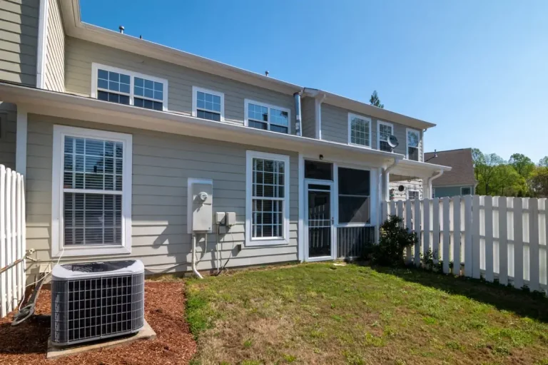 Backyard of a suburban house with air conditioning unit and white picket fence under blue sky
