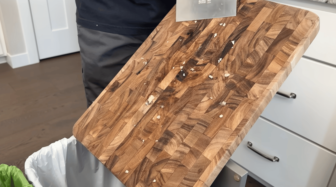wooden cutting board being scraped clean with a metal scraper to remove leftover food residue