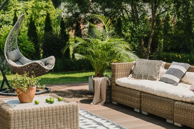 Rattan outdoor furniture and egg chair on wooden deck in sunny garden with lush greenery