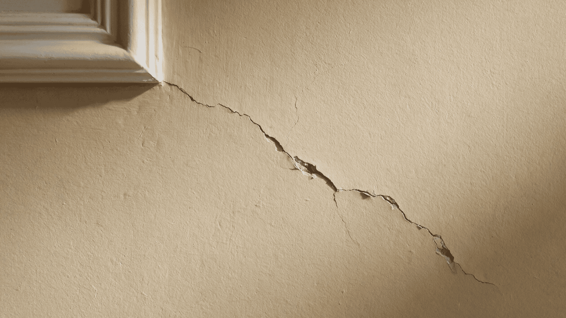 long diagonal crack running across a beige interior wall near white trim with chipped plaster along the split