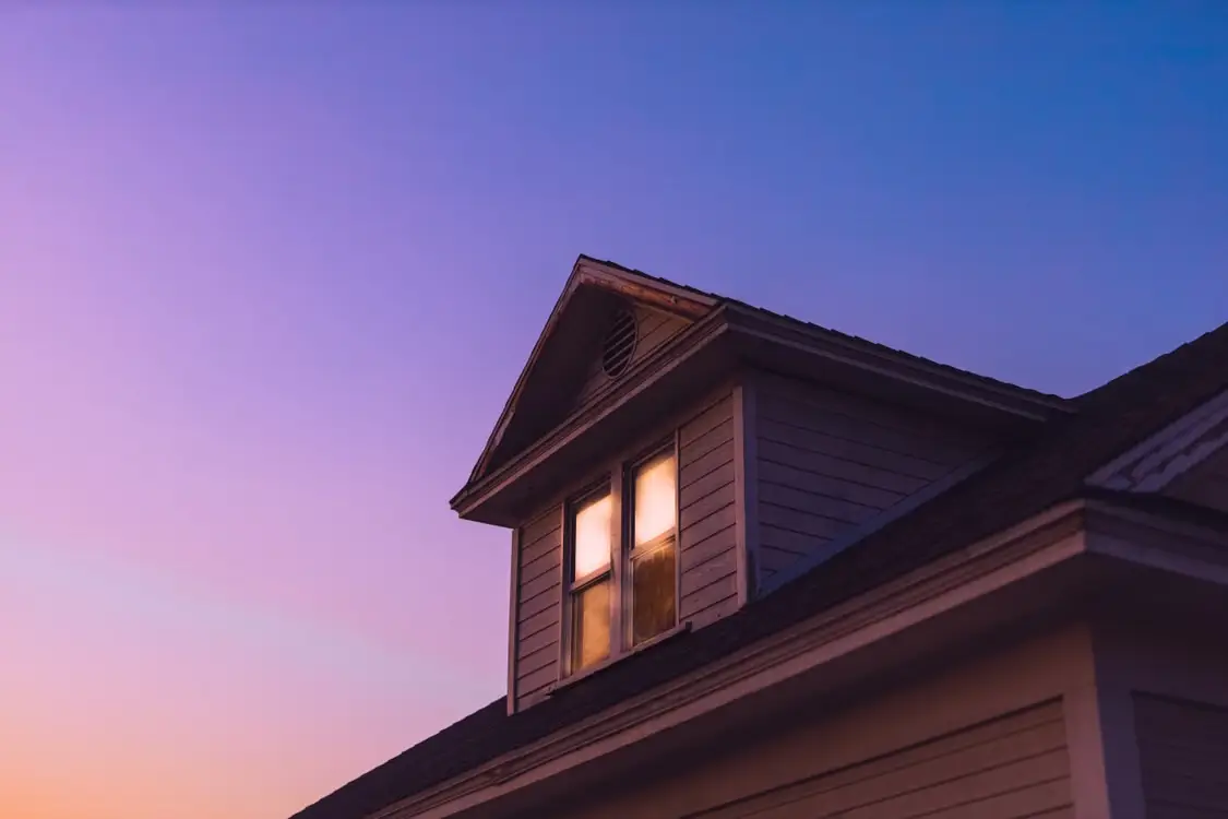 Roof of house with lit window under purple twilight sky