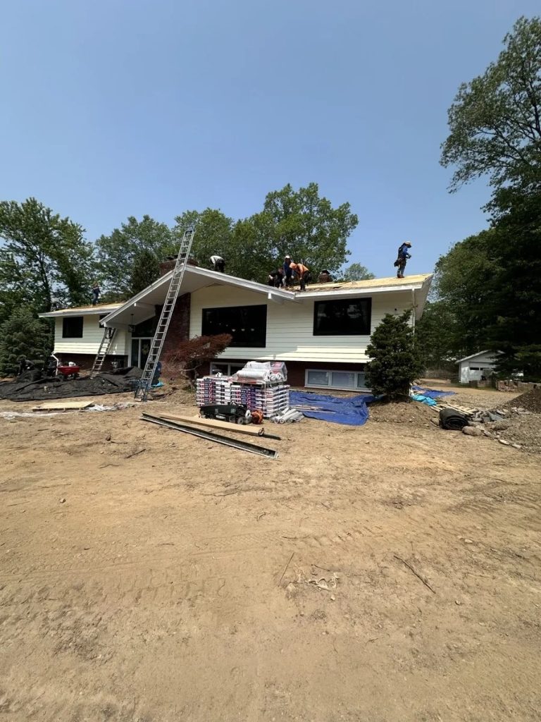 Workers installing shingles on suburban house roof surrounded by trees and construction materials