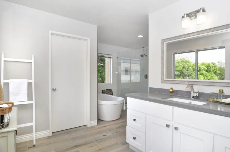 Modern bathroom with white cabinets, gray countertop, freestanding tub, and large window with greenery view