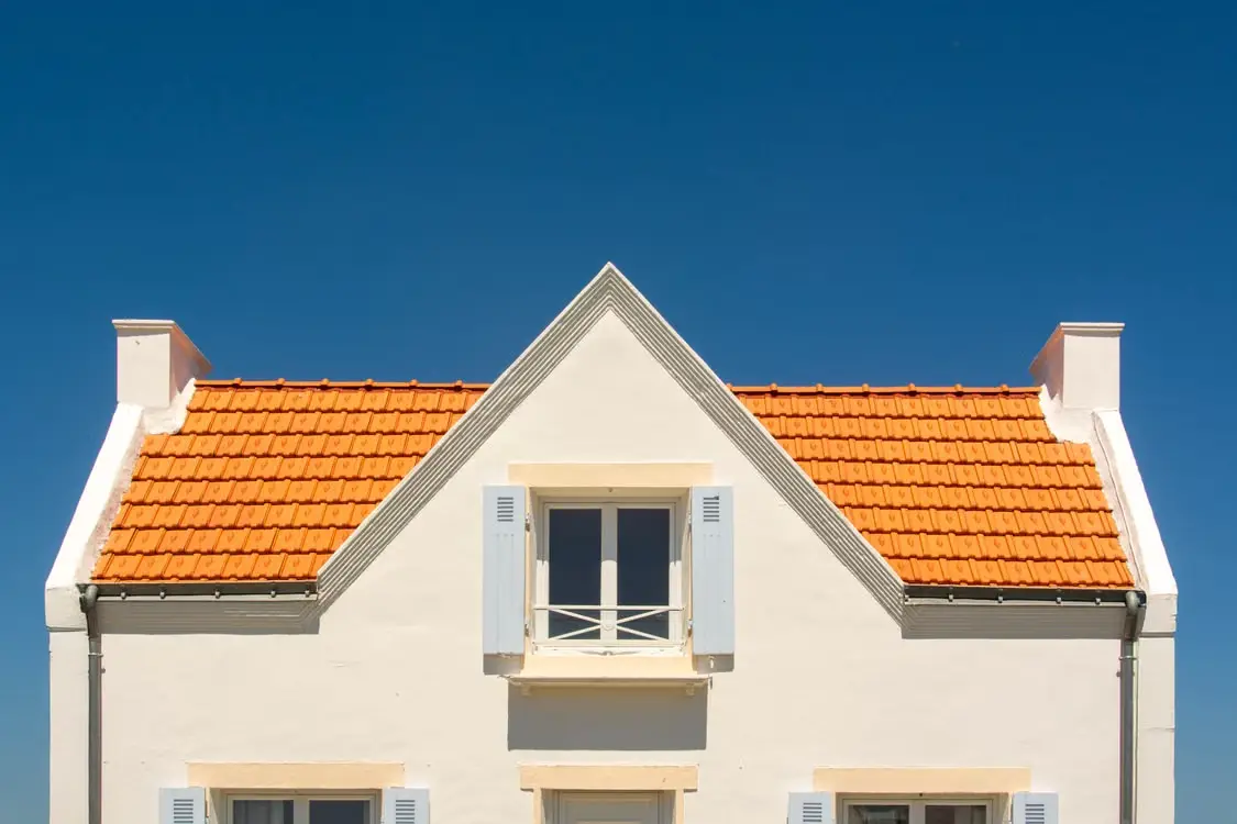 White house facade with orange tiled roof and closed blue shutters under clear blue sky