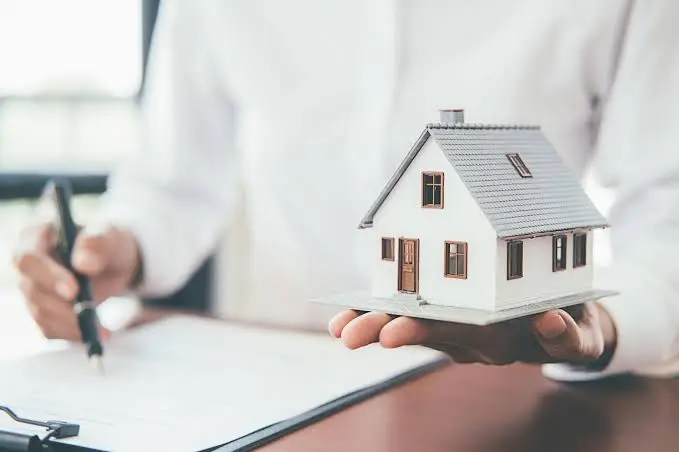 Person holding small model house while signing document in bright office setting