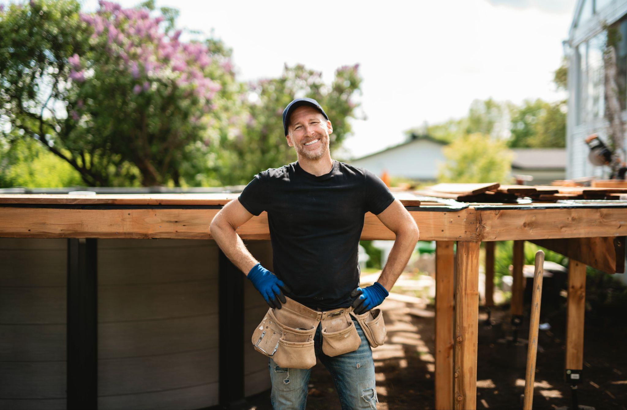 Man in work gloves and tool belt standing by wooden deck under construction outdoors