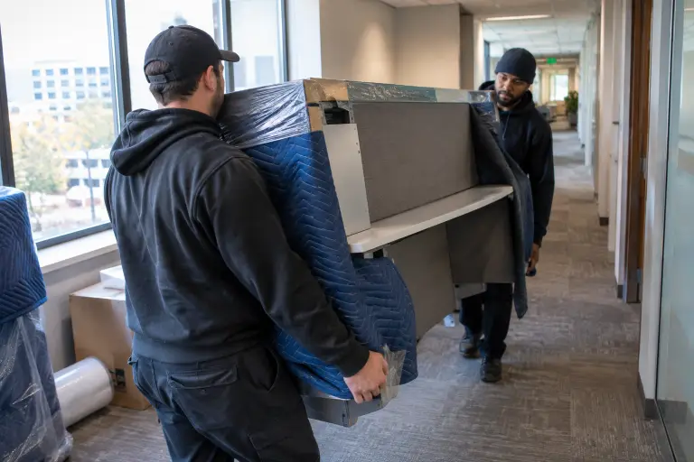 Two workers carrying a wrapped desk through an office hallway with large windows