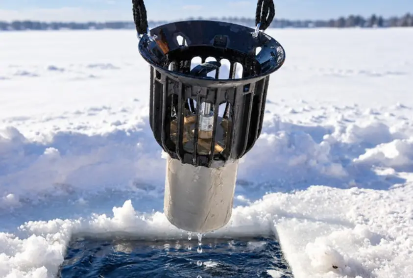 Ice fishing hole with black basket and white container dripping water in snowy landscape